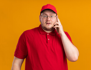 scared overweight young deliveryman in optical glasses talking on phone isolated on orange background with copy space