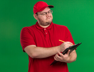 confident overweight young deliveryman in optical glasses writing with pencil on clipboard isolated on green background with copy space