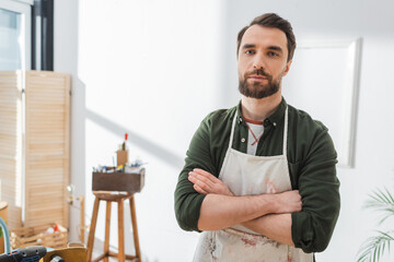 Bearded craftsman in apron crossing arms in workshop.