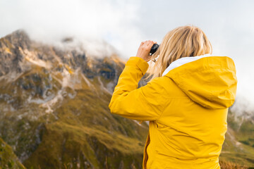 Young blonde woman hiking in mountains in the Giau Pass and looking through binoculars, back view. Wanderlust concept. 