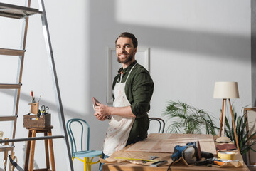 Smiling restorer in apron holding sandpaper near tools and wooden bard in workshop.