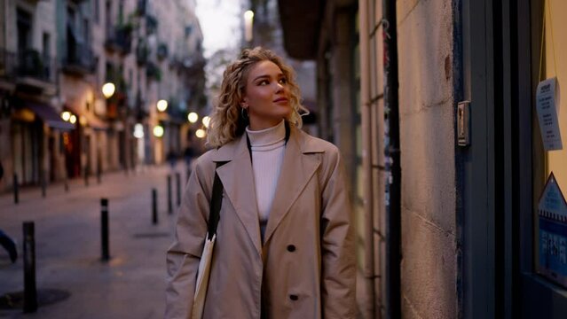 Female tourist enjoys a walk through evening city and stops at shop shopfront window, warm light illuminates her, beautiful street bokeh lights on background. Visiting new places. Slow motion effect