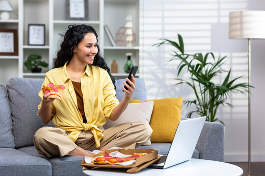 Satisfied woman at home relaxing sitting on sofa in living room and eating pizza, Hispanic woman ordered food online using app on phone, satisfied with fast delivery.