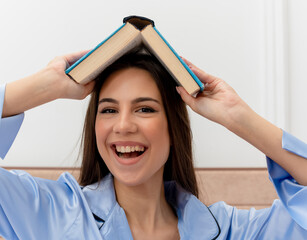 young beautiful woman in blue pajamas sitting on bed holding book over her head looking joyful...