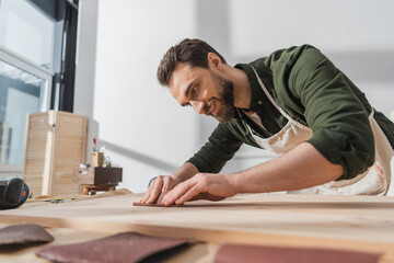 Smiling bearded workman sanding wooden board in workshop.