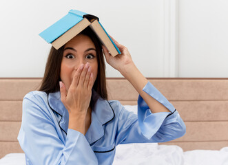 young beautiful woman in blue pajamas sitting on bed holding book over her head looking at camera...