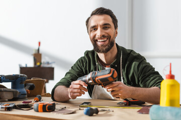 Cheerful carpenter holding electric screwdriver and looking at camera in workshop.