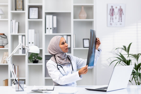 Young successful female doctor in hijab working in clinic inside medical office, muslim woman is angry and thinking x-ray picture sitting at workplace with laptop.