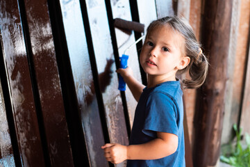 Cute blonde little girl with ponytails carry out painting work, coloring wooden fence border with paint roller and looking at camera in garden outdoors. House working, creative cosmetic reconstruction