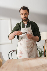Bearded restorer in apron looking at calipers near blurred wooden board.