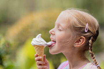 Pretty little caucasian girl with blonde hair eight years old eating licking vanilla ice cream in waffles cone outdoor at hot summer day