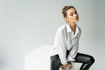 young sensual woman with closed eyes wearing white shirt and black latex pants and sitting on cube...