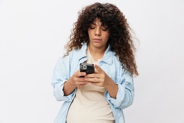 Pregnant woman pensive looking at phone in hand, online communication and correspondence, on white isolated background in t-shirt with blue shirt