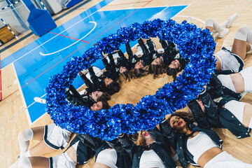 cheerleaders laying on the floor in the circle and holding blue pom-poms up, indoors. High quality photo