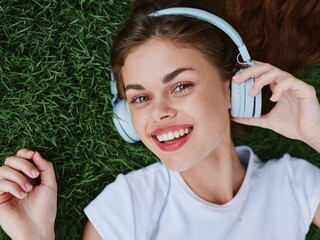 Teenage girl listening to music in headphones lying on the green grass in the park and smiling and laughing in a white T-shirt, spring mood as a lifestyle