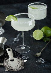 Luxury glasses of Margarita cocktail with fresh limes and bar mat with strainer and jigger, with spoon and ice cubes on black background.