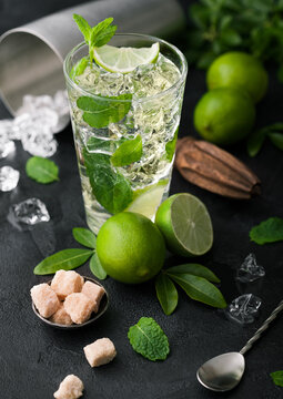 Glass of Mojito cocktail with ice cubes mint and lime on black board with spoon and fresh limes and cane sugar with wooden squeezer and steel shaker with ice.