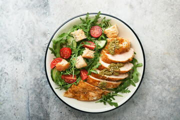 Fresh salad with grilled chicken fillet, arugula, tomatoes, cucumber and pesto sauce, sesame seeds with olive oil in white bowl on light slate background. Healthy lunch menu. Diet food. Top view.