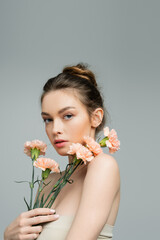 Obraz na płótnie Canvas pretty young woman with natural makeup and bare shoulders looking at camera near peach carnations isolated on grey.