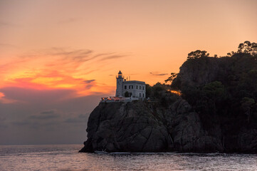 A splendid sunset by the historic lighthouse, Faro di Portofino, at Portofino, Liguria, Italy. A photo taken from the sea below with the rocky mountains and trees in the background.