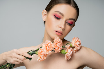 Fototapeta premium pretty woman with pink visage holding fresh carnations near bare shoulder isolated on grey.