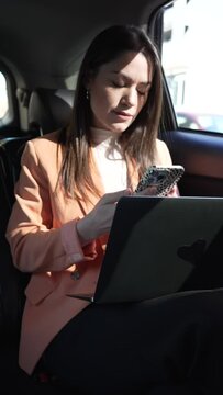 woman inside a car in the back seat, in a cab or car with a driver, writing on her cell phone and working on the computer. Working and doing business on the road