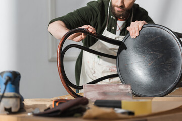 Cropped view of craftsman sanding wooden chair near blurred tools in workshop.