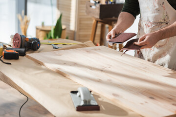 Cropped view of craftsman holding sandpaper near wooden board and tools.