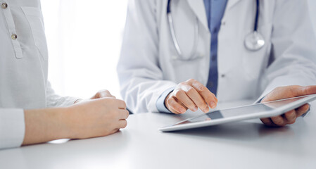 Doctor and patient sitting near each other at the desk in clinic. The focus is on female physician's hands pointing into tablet computer touchpad, close up. Medicine concept