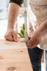 Cropped view of craftsman in dirty apron measuring wooden board with calipers in workshop.
