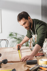 Restorer in apron measuring wooden board with ruler near tools in workshop.