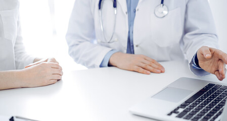 Doctor and patient sitting near each other at the desk in clinic. The focus is on female physician's hands pointing into tablet computer touchpad, close up. Medicine concept