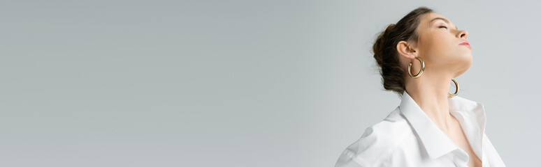 stylish woman in hoop earrings and white shirt posing with closed eyes isolated on grey, banner. © LIGHTFIELD STUDIOS