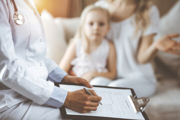 Doctor and patient. Pediatrician using clipboard while examining little girl with her mother at home. Sick and unhappy child at medical exam