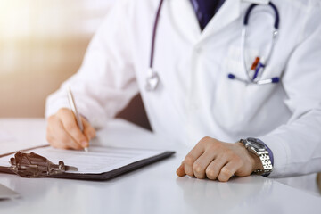 Unknown male doctor sitting and working with clipboard of medication history record in a darkened clinic, glare of light on the background, close-up of hands