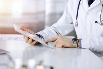 Unknown male doctor sitting and working with tablet computer iin a darkened clinic, glare of light on the background, close-up of hands