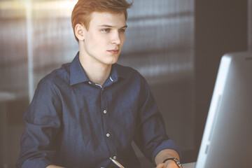 Young blond businessman thinking about strategy at his working place with computer in a darkened office, glare of light on the background