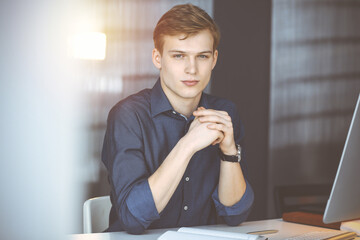 Young blond businessman thinking about strategy at his working place with computer in a darkened...