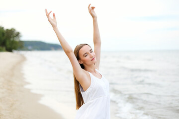 Fototapeta na wymiar Happy smiling woman in free happiness bliss on ocean beach standing with raising hands. Portrait of a multicultural female model in white summer dress enjoying nature 