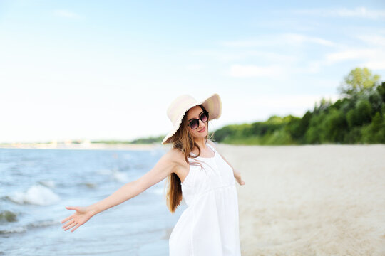Happy smiling woman in free happiness bliss on ocean beach standing with a hat, sunglasses, and open hands. Portrait of a multicultural female model in white summer dress enjoying nature 