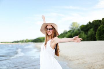 Happy smiling woman in free happiness bliss on ocean beach standing with a hat, sunglasses, and raising hands. Portrait of a multicultural female model in white summer dress enjoying nature 