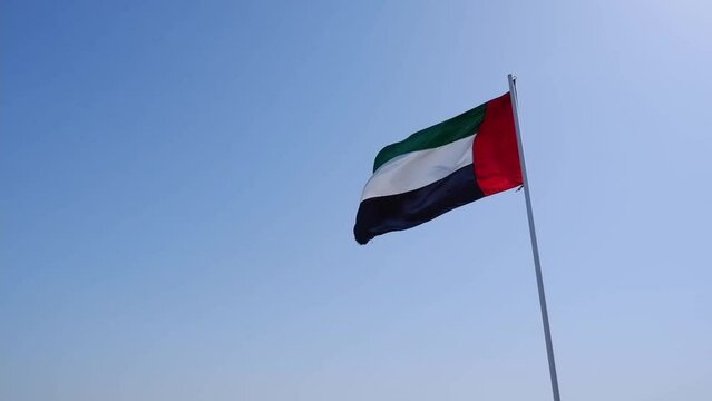The UAE or United Arab Emirates flag flutters in the wind on a flagpole against a blue sky. UAE National Day.