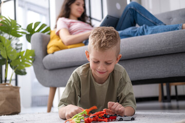 little boy playing on floor dinosaur and cars toys while mother working remotely from home on...