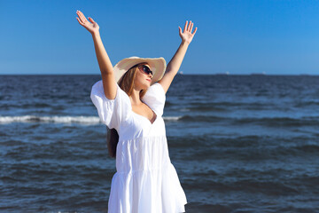 Happy blonde woman is on the ocean beach in a white dress and sunglasses, raising hands