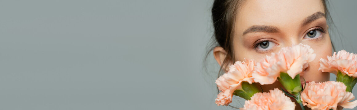 Young Woman Looking At Camera Behind Fresh Carnations Isolated On Grey, Banner.