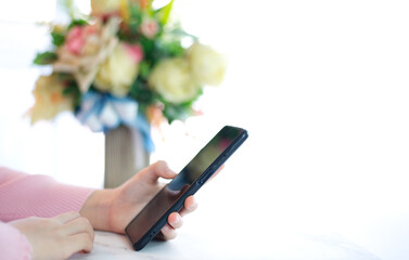 Hands of woman in pink shirt holding smart phone on white table top decorated with bouquet of flowers in a bright room background