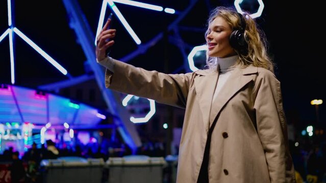 Attractive millennial female in headphones enjoying recreation while taking selfie on cellphone near colorful illuminating ferris wheel. Young woman blogger uses smartphone camera for social media