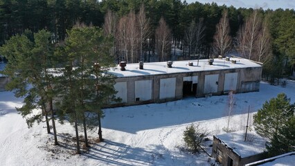 Abandoned building for the storage of military equipment of the former Soviet military base in winter. An old hangar for automotive vehicles under a layer of snow in the middle of a pine forest.