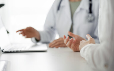 Doctor and patient sitting at the desk in clinic office. The focus is on female physician's hands pointing to laptop computer monitor, close up. Perfect medical service and medicine concept