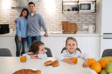Happy family having breakfast on kitchen. Little girl with brother sitting on kitchen and smiling with their parents on background